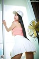 A woman in a white skirt and a pink top posing by a window.